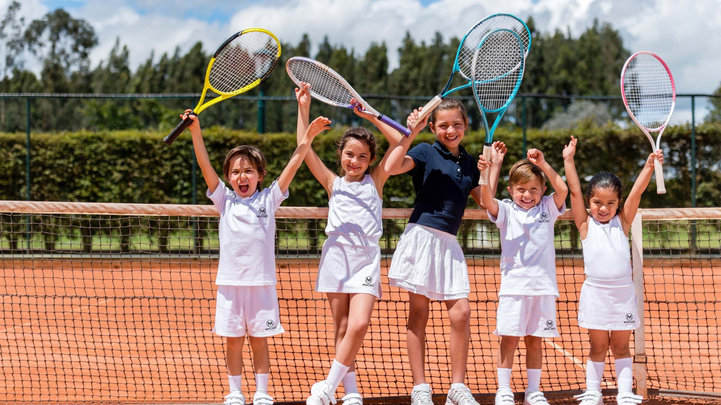 5 garçons et filles heureux portant des shorts, des jupes et des t-shirts Mucho, devant le filet au milieu d'un court de tennis